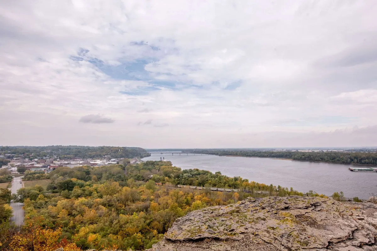 A sweeping view of the Mississippi River, with a prominent bridge, rocky outcrop, and the town nestled along the shoreline, surrounded by vibrant autumn foliage. The image evokes a sense of tranquility and timelessness.