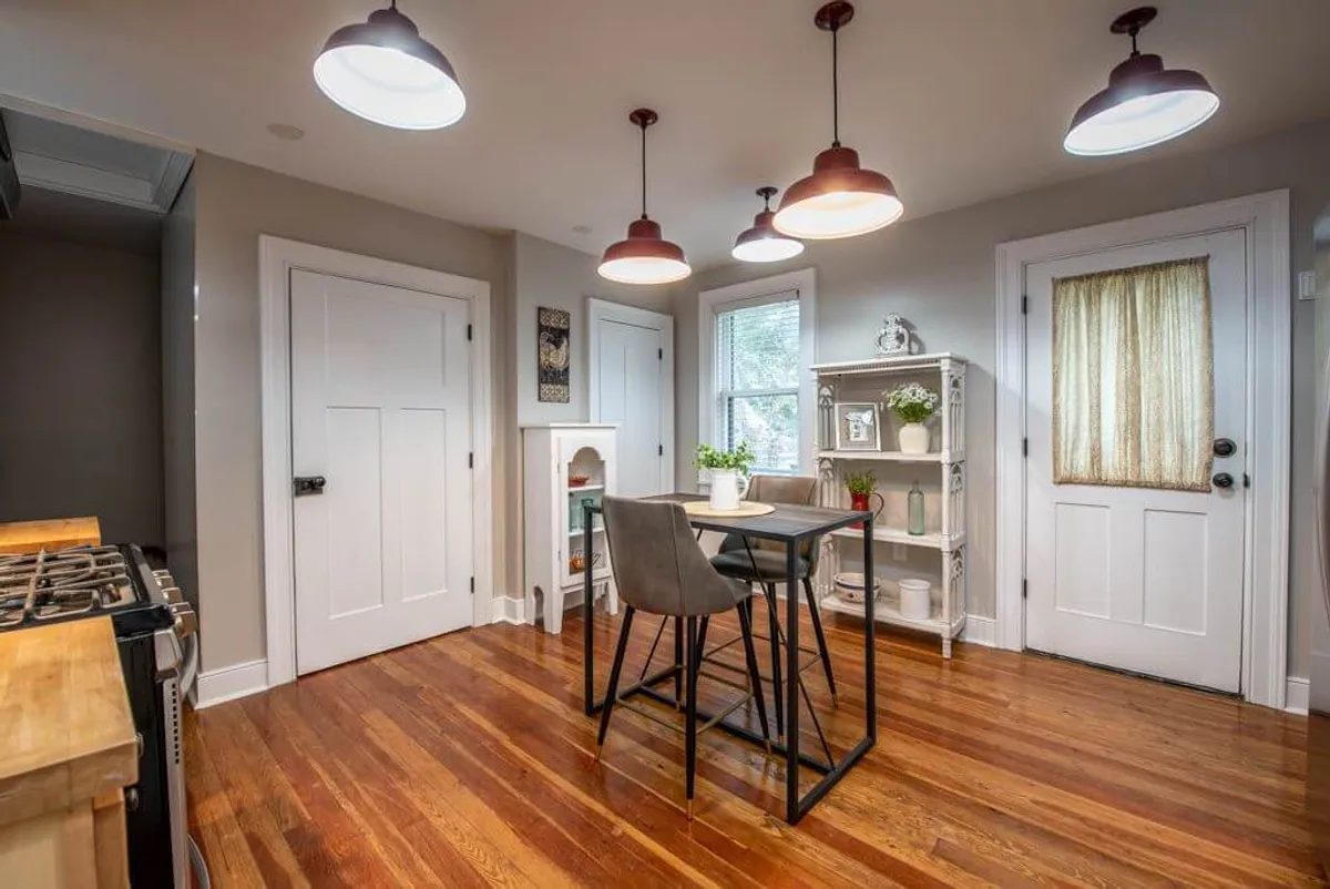 A stylish dining table set under hanging lights, with nearby shelves displaying lush plants, radiates an intimate, inviting ambiance perfect for savoring a candlelit dinner or gathering with loved ones after a day exploring the riverfront.