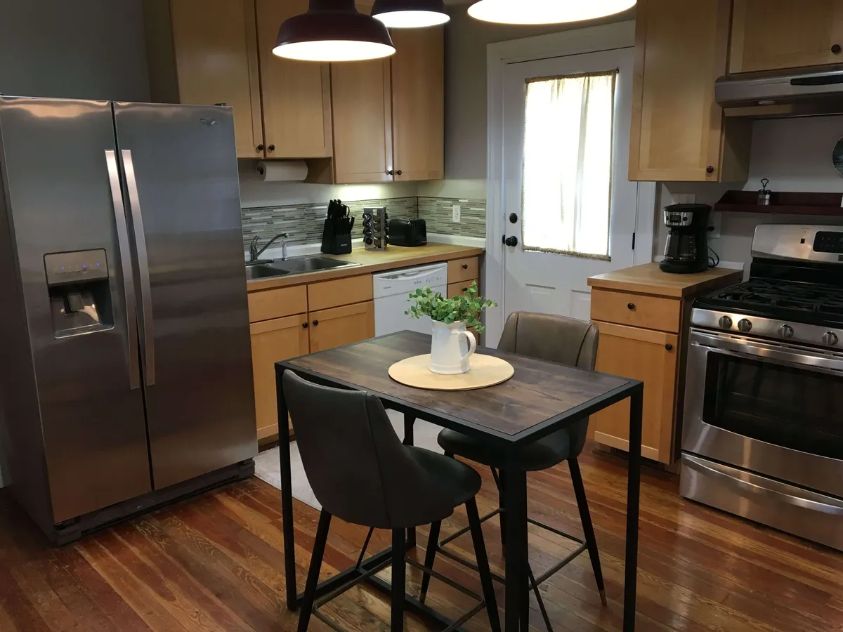 A welcoming kitchen featuring a wooden dining table with a green plant centerpiece, a stainless steel refrigerator, and a homey atmosphere. The space suggests opportunities for casual conversations, coffee breaks, and preparing for outings.