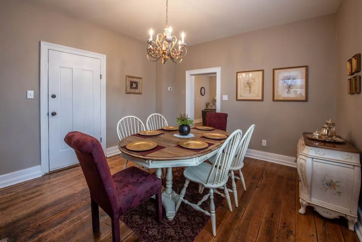 The image showcases a dining area centered on a classic dining table beneath a glowing chandelier, with decorative wall art and a traditional sideboard completing the scene. The mood is set for intimate dinners or lively conversations.