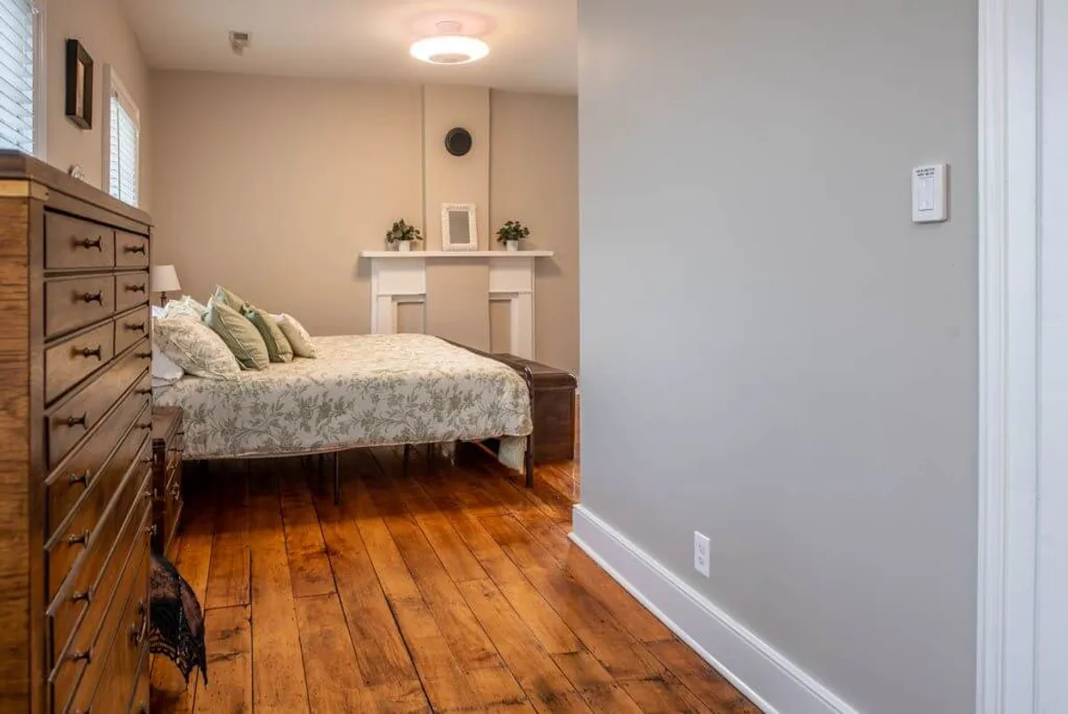 A bedroom with a bed dressed in floral bedding, a wooden dresser, and a cozy fireplace mantel adorned with elegant decor items. The space is bathed in soft light, creating a peaceful and inviting atmosphere ideal for winding down after a long day.