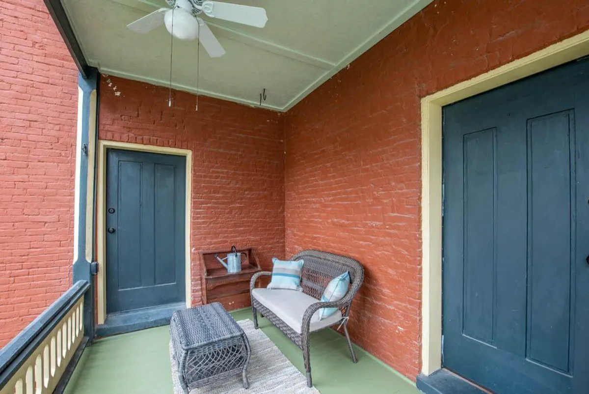 This image features a veranda-like interior space with inviting wicker seating, blue doors, textured walls, and colorful decorative pillows. The setup suggests a breezy, semi-outdoor ambiance ideal for relaxation or casual conversations, evoking the feeling of riverfront leisure.