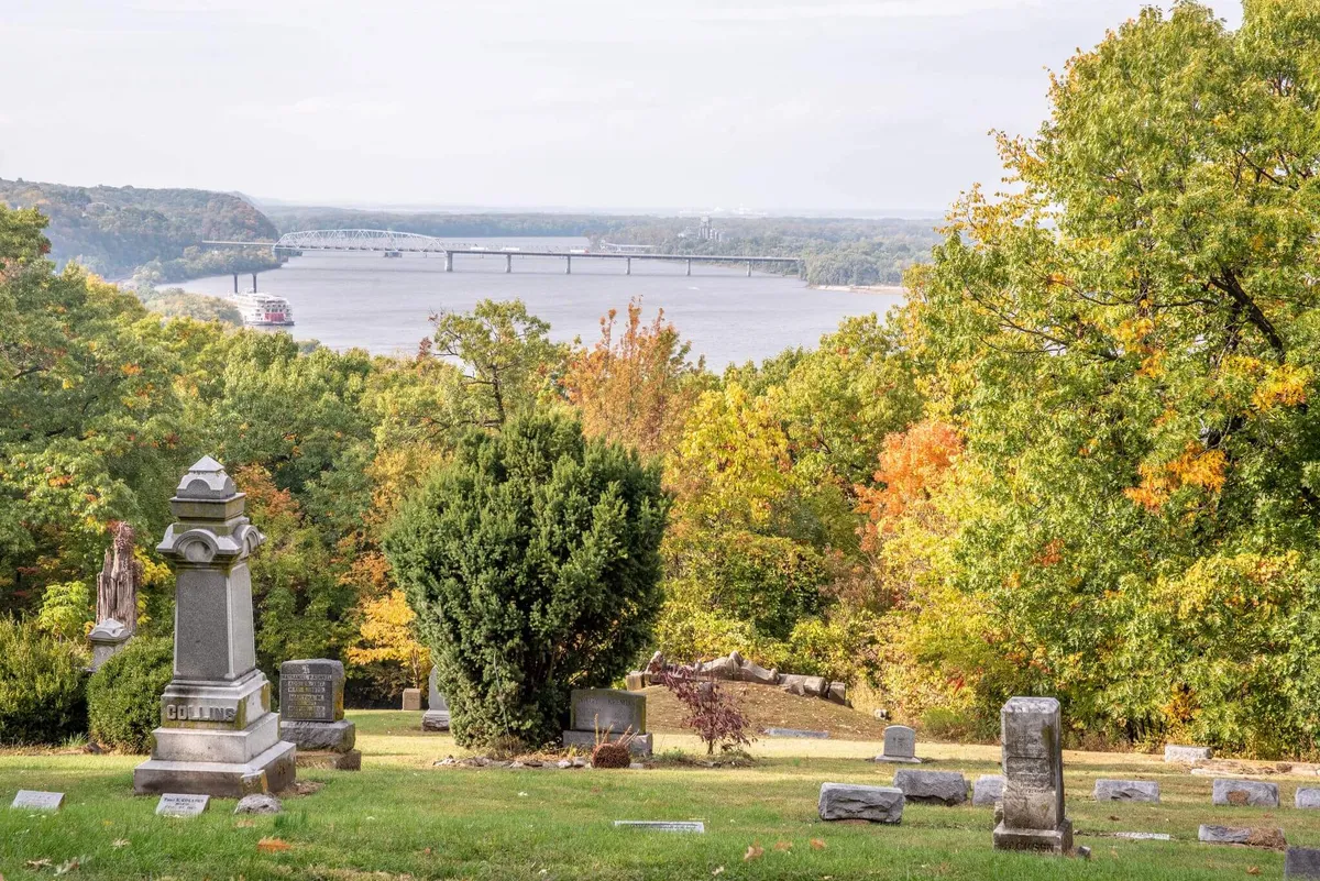 A historic cemetery overlooks the river and bridge, with autumn foliage adding color and atmosphere. Tombstones in the foreground suggest a deep sense of history and reflection, set within a peaceful natural setting.