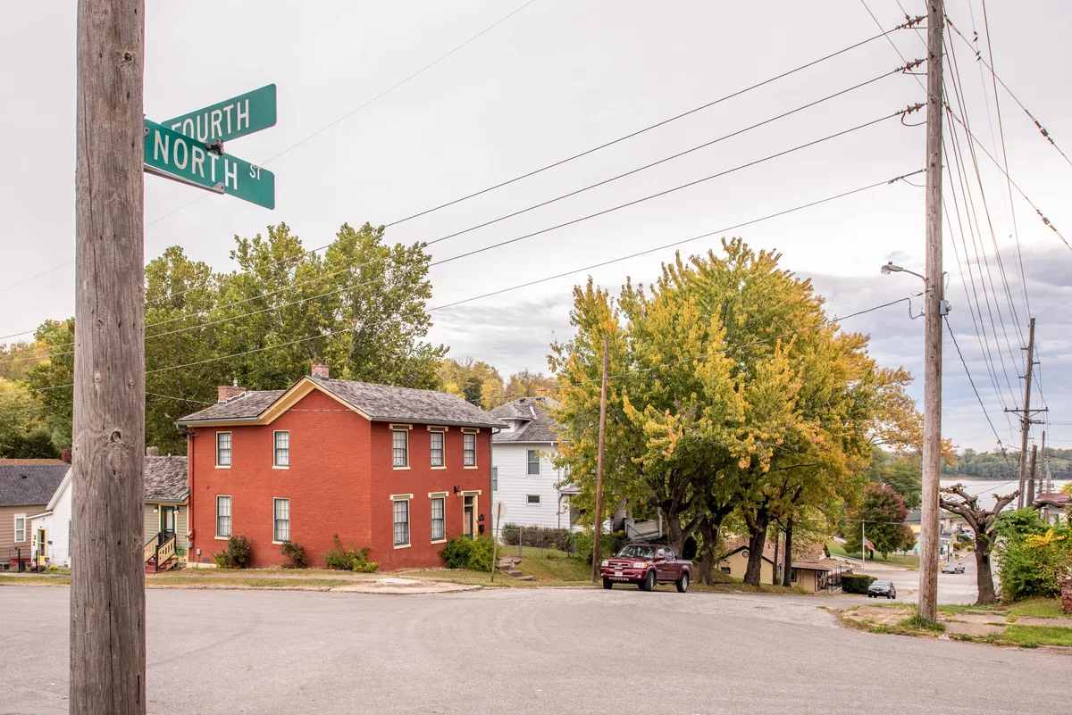 This exterior image captures a quaint red house set at a tree-lined intersection, with surrounding greenery and a nostalgic small-town atmosphere. Power lines and street signs suggest a residential neighborhood that’s walkable and inviting.