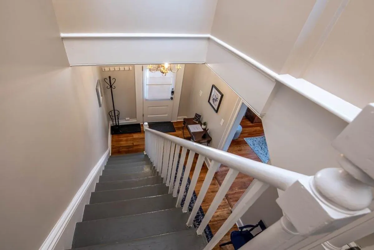 An inviting entryway with a prominent staircase, front door, chandelier, coat rack, and a small table. The layout suggests a welcoming space where guests would pass through, hang coats, and start their journey into town.