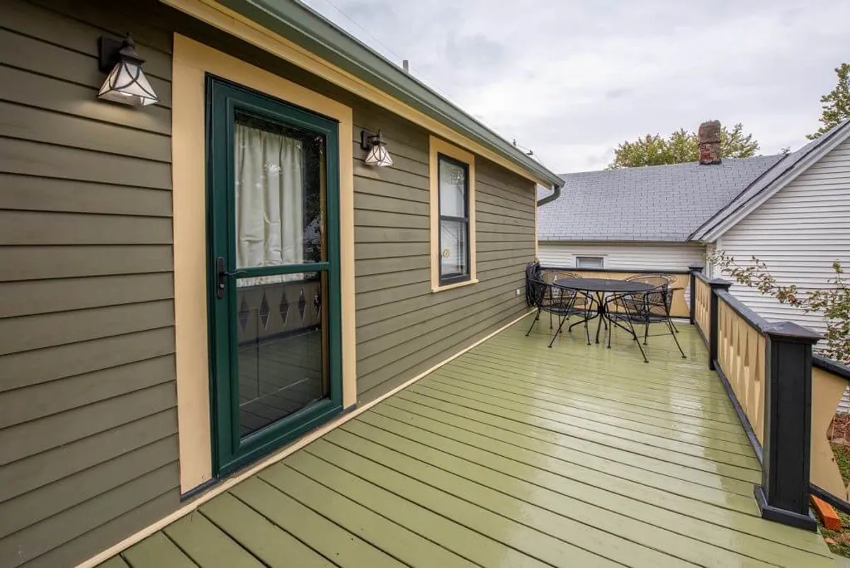 An exterior image showcasing a green door opening onto an outdoor dining set, complemented by decorative light fixtures. The scene radiates a cozy, welcoming mood, perfect for enjoying morning coffee or evening drinks outdoors. The perspective highlights the seamless connection between indoor living and outdoor river views.